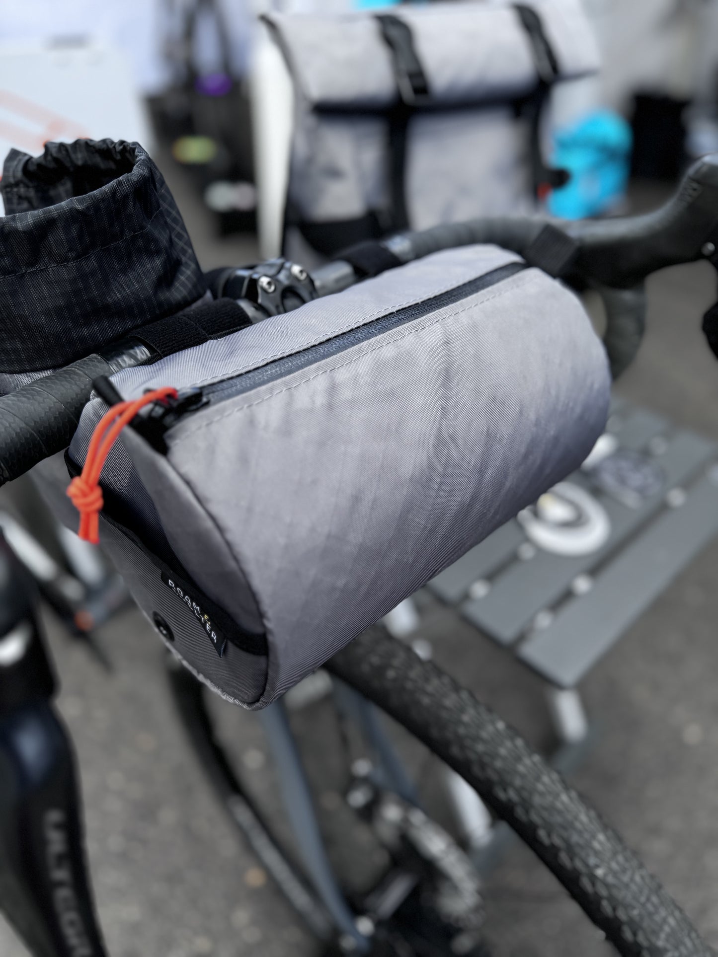 A gray Round Bar Bag attached to a bicycle's handlebar, featuring a YKK AquaGuard zipper and a Velcro strap.
