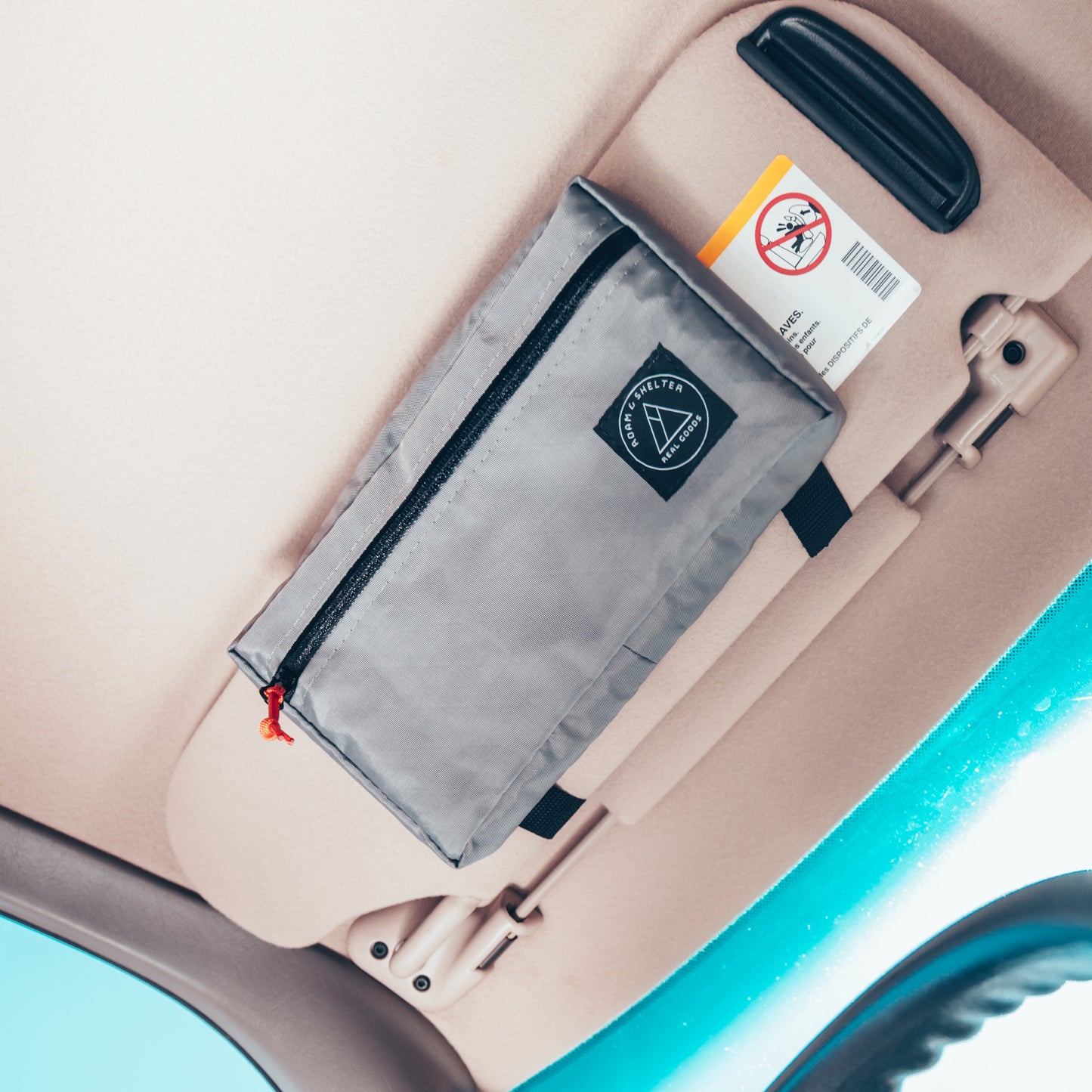 A gray visor bag attached to the interior visor of a vehicle, featuring a horizontal front zip opening and an orange zipper pull. Handmade in Portland Oregon USA.