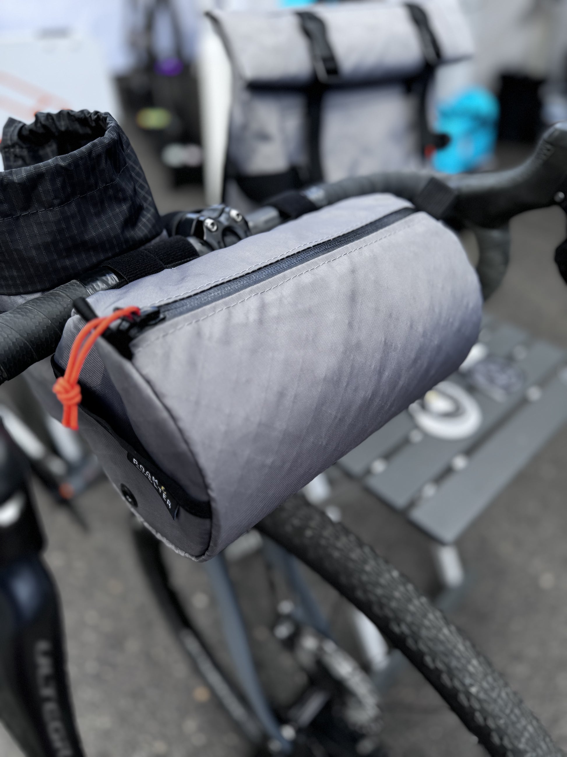 A gray Round Bar Bag attached to a bicycle's handlebar, featuring a YKK AquaGuard zipper and a Velcro strap.
